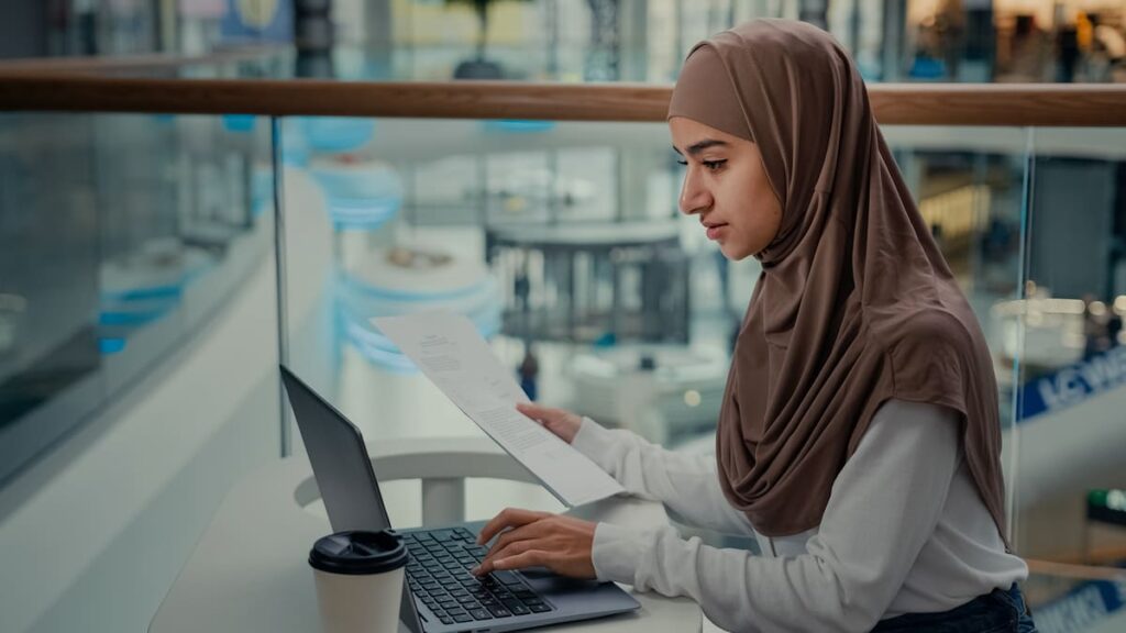 Mujer con hiyab trabajando con un ordenador portátil mientras revisa documentos, en un entorno profesional relacionado con la inclusión laboral y las migraciones.