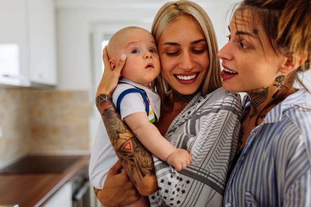 Dos mujeres sonrientes junto a su bebé en el hogar, representando la diversidad familiar y las familias homoparentales.